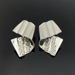 Puede incluir: Un par de pendientes plateados con un diseño de metal esculpido y plegado. Cada pendiente tiene una sección superior lisa y curva, y una parte inferior con un patrón detallado de eslabones de cadena en relieve con acabado metálico.