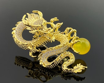 Puede incluir: Broche de dragón dorado con detalles intrincados. El dragón está adornado con cristales transparentes y sostiene un orbe amarillo. La broche se encuentra sobre una superficie negra reflectante.