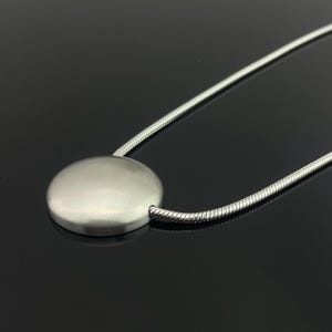 以下が含まれることがあります： つや消しシルバーのペンダントネックレス。ペンダントは、シルバーのスネークチェーンに取り付けられた平らな円盤です。ネックレスは、暗く反射する表面に表示されています。