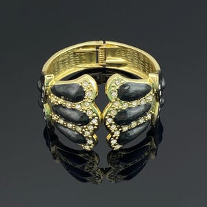 May include: A gold-tone hinged cuff bracelet featuring a symmetrical design with black enamel teardrop-shaped inlays and clear crystal accents. The interior of the metal band has a textured finish and a secure clasp mechanism for closure.