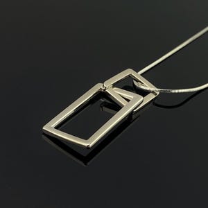 May include: A silver-toned necklace featuring two interlocking rectangular frames as a pendant. The pendant is suspended from a thin, flexible snake chain. The metal has a smooth, polished finish that reflects light, creating a clean and geometric look.