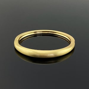 Puede incluir: Un brazalete de color dorado con un acabado mate. El brazalete es una banda sólida con un exterior liso y redondeado. La joya se exhibe sobre una superficie negra reflectante.