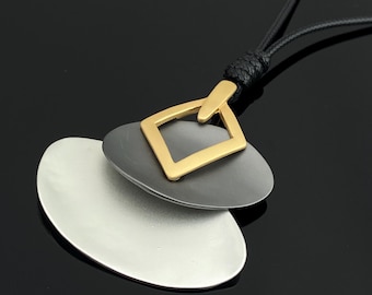 May include: A pendant necklace featuring a black cord and three layered geometric shapes. The top element is a gold-toned square, the middle is a dark gray oval, and the bottom is a larger silver-toned oval. The pieces are connected by a knot.
