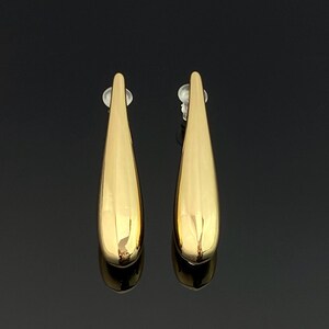 May include: A pair of gold-toned drop earrings featuring a smooth, elongated teardrop shape with a polished, reflective surface. Each earring is attached to a clear plastic post backing, showcasing a simple, solid metal design with no additional embellishments.
