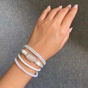Könnte beinhalten: Ein Set aus vier geschichteten Armbändern, bestehend aus einem weißen Perlenstrang, einer weißen geflochtenen Kordel, einem weißen Riemen mit einem ovalen Stein in silberfarbener Fassung und einem dünnen silberfarbenen Band mit kleinen klaren Kristallen.