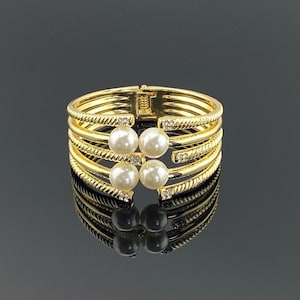 May include: Gold-toned bracelet with four rows, each with a textured design. The bracelet features four faux pearls and small clear crystals. The bracelet is reflected in the glossy black surface.