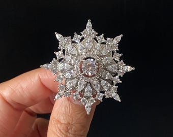Könnte beinhalten: Eine silberfarbene Brosche in Form einer Schneeflocke mit einem zentralen runden Stein, umgeben von mehreren Schichten aus birnenförmigen und marquise-geschliffenen klaren Kristallen, die in einem strahlenförmigen Muster angeordnet sind.