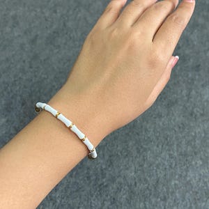 Op de afbeelding: Een armband bestaande uit een reeks gladde witte cilindrische segmenten, gescheiden door kleine, gepolijste goudkleurige metalen ringen. Het ontwerp bootst het uiterlijk van bamboestengels na en vormt een herhalend patroon rond de pols.