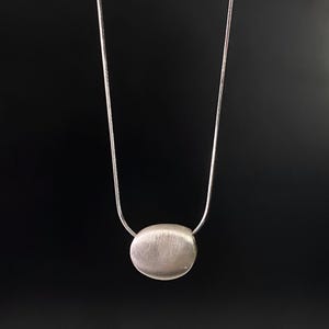 Könnte beinhalten: Silberfarbene Halskette mit gebürstetem, ovalem Anhänger. Der Anhänger hängt an einer zarten Silberkette vor schwarzem Hintergrund. Der Schmuck ist minimalistisch.