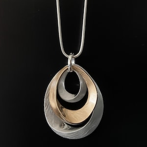 May include: A pendant necklace featuring three nested, teardrop-shaped loops. The outer and inner loops have a brushed silver finish, while the middle loop has a brushed gold finish. The pendant hangs from a thin, silver-toned snake chain.