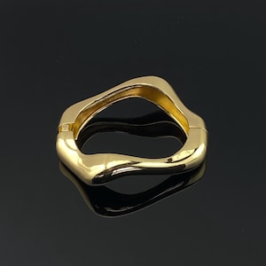 May include: A gold-colored, wavy-shaped bracelet with a hinged clasp. The bracelet has a smooth, polished surface and reflects light, creating a shiny appearance. The design is modern and abstract, with a flowing, organic form.