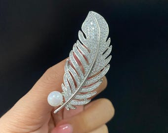 May include: A silver-toned feather brooch encrusted with numerous small, clear, sparkling stones. The feather design features a central stem and a single, smooth, round white pearl accent positioned at the base of the quill, adding contrast.