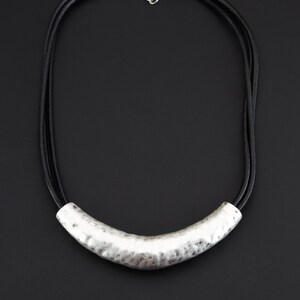 May include: A necklace featuring a curved, hammered silver-tone metal tube pendant suspended from three thin black cords. The necklace includes a silver-tone lobster claw clasp and an adjustable chain link extender at the back for secure closure.