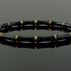 May include: A black and gold-coloured bamboo-style bracelet. The bracelet has black segments with gold-coloured accents at the joints. The jewellery is displayed on a reflective surface, highlighting its design.