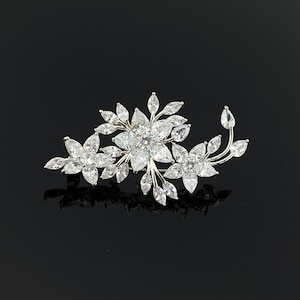 Puede incluir: Broche floral plateado con tres grupos de flores y detalles de hojas. La broche está adornada con numerosos cristales claros y brillantes, creando un efecto deslumbrante. El diseño es simétrico y elegante, adecuado para eventos formales.