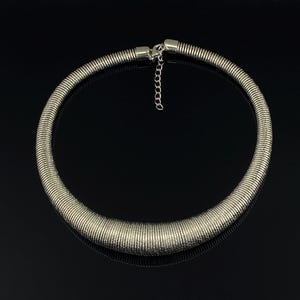 Peut inclure: Un collier ras-de-cou en métal argenté composé d'une chaîne serpent épaisse et flexible. Le collier présente un profil arrondi et lisse qui s'affine légèrement vers l'arrière, où il se ferme par un fermoir mousqueton et une chaînette d'extension.