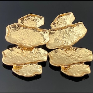 May include: Four gold-colored earrings with a hammered texture. Each earring has an irregular, organic shape, with a larger, flat base and a smaller, raised top section. The earrings are displayed against a dark background, highlighting their metallic sheen.