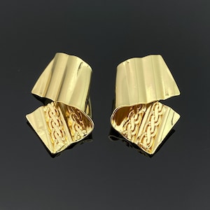 Può includere: Un paio di orecchini color oro caratterizzati da una sezione superiore liscia e curva e una sezione inferiore testurizzata con un intricato motivo a catena intrecciata. Gli orecchini presentano una finitura lucida e una forma scultorea.