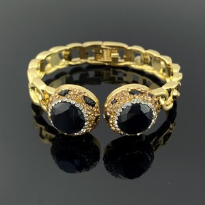 Puede incluir: Una pulsera de brazalete de metal dorado con una banda de eslabones y dos puntos focales circulares. Cada elemento circular contiene una gran piedra negra facetada rodeada por un borde de cristales más pequeños, transparentes y de color champán.