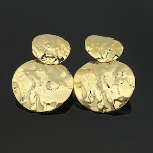 Puede incluir: Un par de pendientes dorados. Cada pendiente presenta un diseño texturizado y martillado con una pequeña pieza superior irregular y una pieza inferior redonda más grande. Los pendientes tienen una superficie brillante y reflectante.