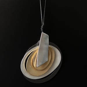 May include: A silver and gold-toned pendant necklace with an abstract design. The pendant features layered oval shapes and a rectangular element, suspended from a black cord. The necklace is set against a dark background.
