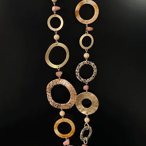 Può includere: Una lunga collana con una varietà di elementi dorati e rosa. La collana presenta grandi pendenti circolari, anelli più piccoli e perline di pietra rosa. Lo sfondo è nero.
