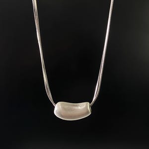 Könnte beinhalten: Eine silberne Halskette mit einem gebogenen, rechteckigen Anhänger. Der Anhänger hat eine helle, matte Silberfarbe und hängt an einer zarten, mehrsträngigen Kette. Der Hintergrund ist einfarbig schwarz.