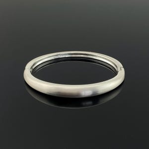 May include: A silver-coloured, hinged bangle bracelet with a brushed metal finish. The bracelet has a smooth, rounded design and a secure clasp. The jewellery is displayed on a reflective black surface.
