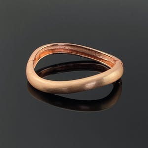 Puede incluir: Un brazalete de color oro rosa con un diseño curvo único. El brazalete tiene un acabado liso y mate y un cierre de bisagra. El brazalete se muestra sobre una superficie negra reflectante.