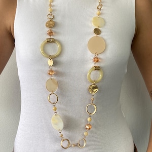Puede incluir: Un collar largo, dorado, con una variedad de elementos decorativos. El collar presenta colgantes grandes, redondos y ovalados de color crema, anillos dorados y pequeñas cuentas de color ámbar. El collar se lleva sobre una camiseta sin mangas blanca.