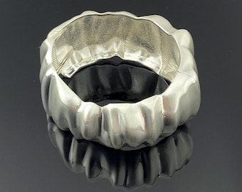 May include: A wide, polished silver-tone cuff bracelet featuring a continuous, undulating, and organic rippled texture around the entire exterior surface. The bracelet has a hinged opening mechanism visible on the side, creating a circular shape.