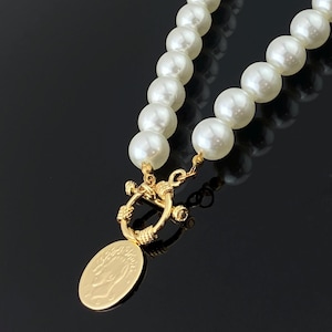 Puede incluir: Un collar que presenta una hilera de cuentas esféricas blancas conectadas a un cierre de palanca en tono dorado. Un colgante circular dorado con el perfil de una cabeza en relieve está unido al cierre, creando un punto focal decorativo.