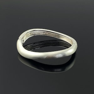 Pode incluir: Uma pulseira de bracelete prateada com um design oval curvo. A pulseira tem um acabamento em metal escovado e um fecho articulado. A pulseira é mostrada em uma superfície preta reflexiva.