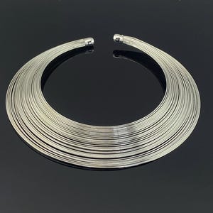 以下が含まれることがあります： シルバーカラーのチョーカーネックレス。 ネックレスは、多数の細い平行な金属線で構成されており、幅広のオープンチョーカーデザインを形成しています。 ネックレスの両端は丸みを帯びたキャップ付きの端子です。 黒い表面に表示されています。