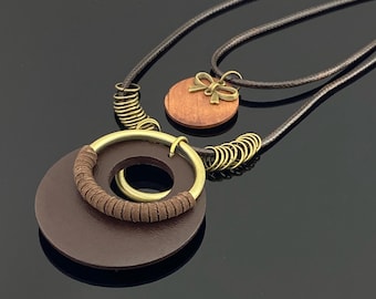 May include: A layered necklace featuring a dark brown cord with a large dark brown circular pendant, a brass ring wrapped in brown leather, and a smaller wooden circular pendant adorned with a small brass bow charm and brass rings.