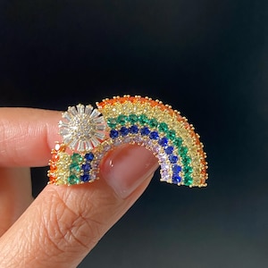 Könnte beinhalten: Eine goldfarbene Brosche in Regenbogenform mit Reihen kleiner, mehrfarbiger Edelsteine in Orange, Gelb, Grün, Blau und Violett. Ein kreisförmiges, sonnenartiges Element mit klaren, länglichen Steinen ist an der linken Seite befestigt.
