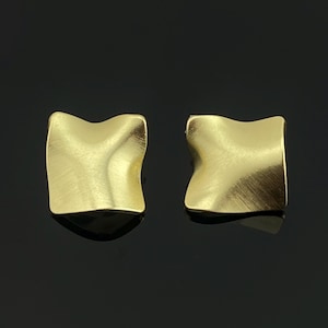 May include: A pair of gold-toned stud earrings featuring a square shape with a soft, wavy, and undulating surface texture. The metal has a brushed, matte finish that catches the light across the curved, concave contours of each individual earring.