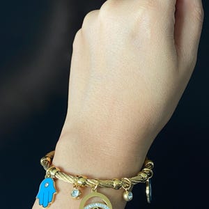Puede incluir: Pulsera dorada con múltiples colgantes. La pulsera presenta un colgante de mano de Fátima azul, un colgante circular con un diseño de ojo y varios colgantes pequeños colgantes. La pulsera se lleva en la muñeca.