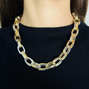 May include: A chunky chain necklace featuring alternating oval links in polished gold and silver tones. The necklace sits at the base of the neck and is composed of thick, interlocking metal links that create a bold, textured appearance.