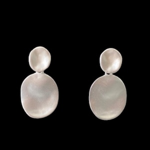 May include: A pair of silver-toned drop earrings featuring a small circular top disc connected to a larger, slightly irregular oval-shaped bottom disc. The metal surface has a soft, matte finish with subtle indentations and a smooth, reflective texture.