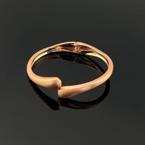 Puede incluir: Un brazalete articulado de oro rosa. El brazalete tiene un acabado mate y un cierre de broche. El diseño es simple y elegante, adecuado para el uso diario o ocasiones especiales. El brazalete se muestra sobre un fondo oscuro.