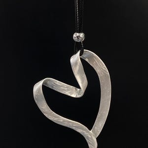 Può includere: Una collana con pendente a forma di cuore argentato. Il pendente a forma di cuore è realizzato in metallo spazzolato e strutturato e pende da un cordino nero. Una piccola perla d'argento si trova nella parte superiore del pendente. Lo sfondo è nero.