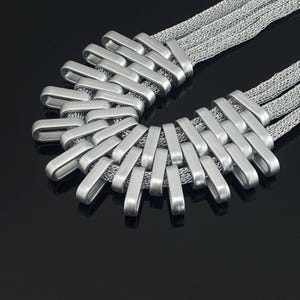 Op de afbeelding: Een zilverkleurige ketting met een reeks onderling verbonden, gladde, rechthoekige metalen lussen in een gebogen, waaiervormig patroon. De lussen zijn geregen aan meerdere strengen van een fijne, flexibele zilverkleurige metalen gaasketting.