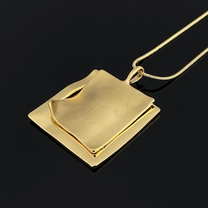 Puede incluir: Un collar con colgante en tono dorado que presenta un diseño cuadrado en capas. El colgante consta de una base cuadrada plana rematada con una pieza cuadrada más pequeña y ligeramente curvada con una superficie lisa y reflectante. Cuelga de una cadena fina.