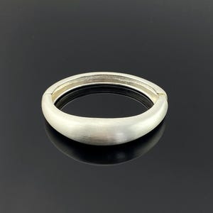 Puede incluir: Un brazalete articulado de color plateado. El brazalete tiene un acabado de metal cepillado liso y una forma ovalada redondeada. El cierre es visible en el lateral. El brazalete se muestra sobre una superficie negra.
