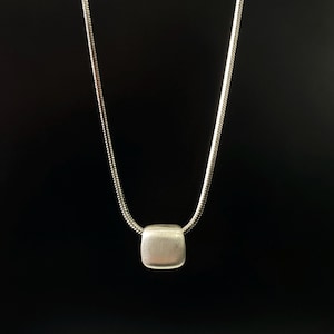 May include: A silver-toned necklace with a square pendant. The pendant is a rounded square shape and hangs from a thin, snake chain. The background is solid black.