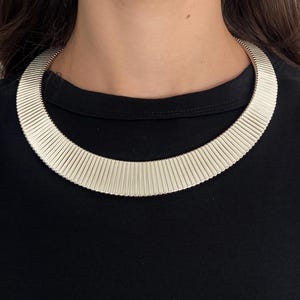 Könnte beinhalten: Eine silberfarbene Halskette in Form eines breiten, starren Reifs, der aus zahlreichen dünnen, vertikalen und parallelen Metallsegmenten besteht. Die Kette hat eine glatte, polierte Oberfläche und liegt eng und geschwungen am Hals an.