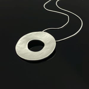 Puede incluir: Collar plateado con un colgante circular grande y texturizado. El colgante tiene un aspecto martillado y una abertura central. La cadena es una fina cadena de serpiente plateada. La joya se muestra sobre un fondo oscuro.