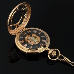 May include: Gold-tone pocket watch with an open face and a black dial featuring Roman numerals. The watch has a detailed, visible inner mechanism and a matching chain. The watch case is open, revealing the intricate design.