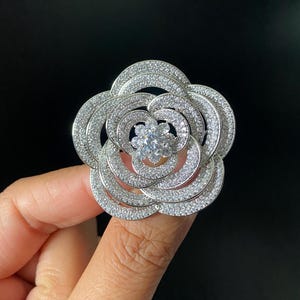 Puede incluir: Un broche de tono plateado con forma de flor en floración con cinco pétalos superpuestos. Toda la superficie de los pétalos está incrustada con pequeñas piedras claras y brillantes, y una piedra clara más grande está engastada en el centro.